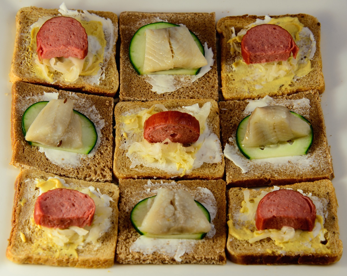 A plate of open face Danish-style sandwiches, as seen from above, arranged in a checkerboard pattern. The sausages are cut into half-round coins, and the herring is cut into trapezoids.